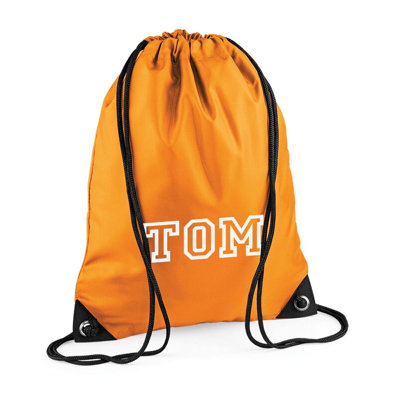 May include: Orange drawstring bag with the name "TOM" printed in white block letters. The bag has black drawstrings and black reinforced corners. The bag is made of a smooth, possibly nylon, material.