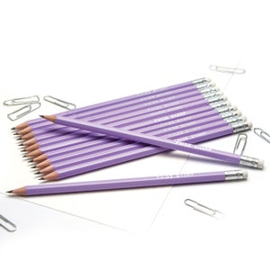 Puede incluir: Un paquete de 15 lápices de color lavanda con detalles plateados y texto blanco que dice "Paige Sharp" en cada lápiz. Los lápices están dispuestos en forma de abanico sobre una superficie blanca con clips de papel plateados esparcidos alrededor.