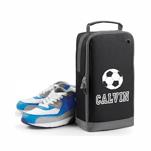 以下が含まれることがあります： 白いサッカーボールと「CALVIN」という名前が印刷された黒い靴袋。青い靴と白い靴のペアが袋の隣にあります。