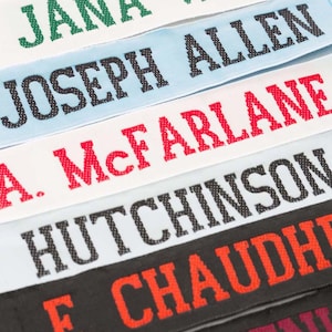 May include: Close-up of several personalised name tapes in various colours. The names are embroidered in bold, capital letters. The tapes are in green, light blue, white, grey, black, and burgundy.