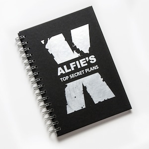 Op de afbeelding: Een zwart spiraalnotitieblok met een zilveren folieontwerp van een letter X en de tekst "Alfie's Top Secret Plans".