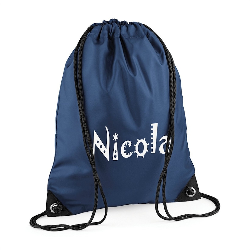 May include: Navy blue drawstring backpack with black drawstrings and grommets. The name "Nicola" is printed in white, with decorative embellishments on the letters. The bag is made of a smooth, durable material.