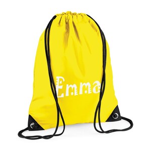 May include: A bright yellow drawstring backpack with black drawstrings and eyelets. The name "Emma" is printed in white, stylized lettering on the front of the bag. The bag is made of a lightweight material.