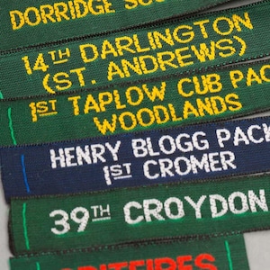 Pode incluir: Close-up de várias fitas de tecido com texto bordado. As fitas são de várias cores, incluindo verde e azul marinho. O texto inclui "DORRIDGE", "14TH DARLINGTON", "HENRY BLOGG PACK" e "39TH CROYDON".