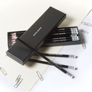 May include: A black box with the text "AUSTIN DRAPER" on it, containing a set of black pencils with white erasers. The pencils are labeled with the text "AUSTIN DRAPER".