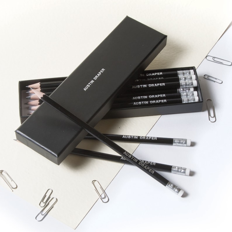 May include: A black box with the text "AUSTIN DRAPER" on it, containing a set of black pencils with white erasers. The pencils are labeled with the text "AUSTIN DRAPER".