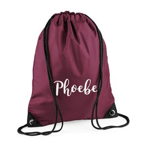 May include: A burgundy drawstring backpack with black drawstrings and grommets. The name "Phoebe" is printed in a white script font across the front of the bag. The bag is made of a smooth, possibly nylon, material.