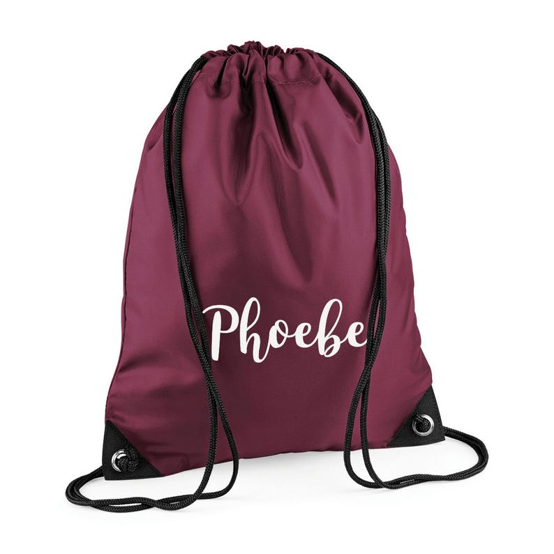 May include: A burgundy drawstring backpack with black drawstrings and grommets. The name "Phoebe" is printed in a white script font across the front of the bag. The bag is made of a smooth, possibly nylon, material.