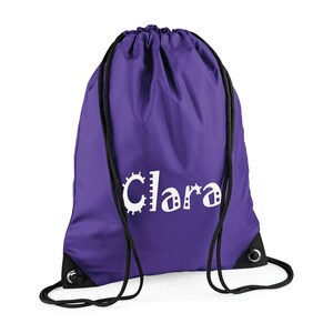 May include: A purple drawstring backpack with black drawstrings and grommets. The name "Clara" is printed in white, with a unique, playful font. The bag is made of a smooth, durable material, perfect for carrying items.
