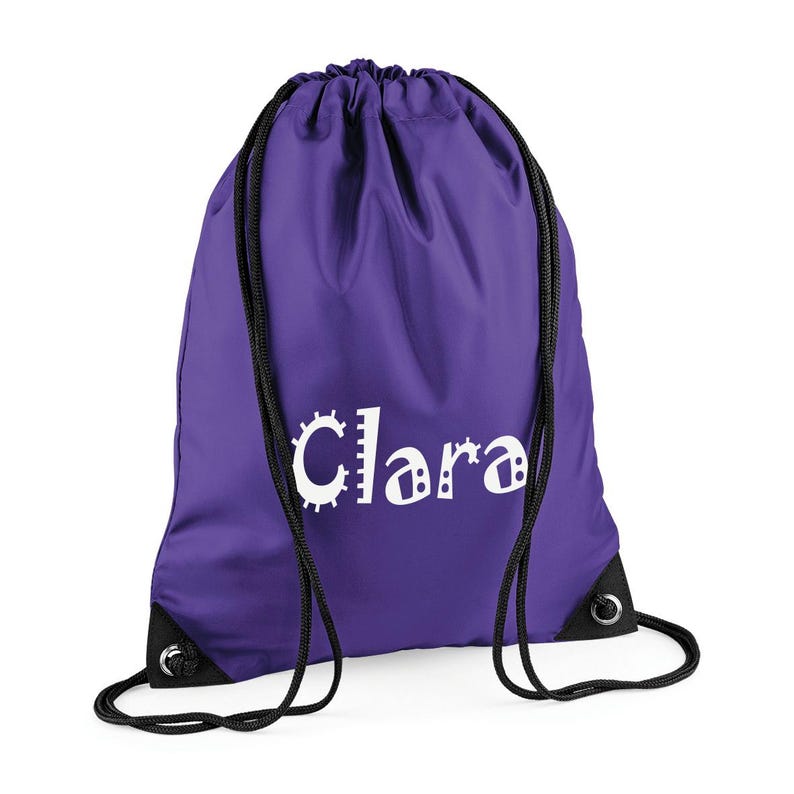 May include: A purple drawstring backpack with black drawstrings and grommets. The name "Clara" is printed in white, with a unique, playful font. The bag is made of a smooth, durable material, perfect for carrying items.
