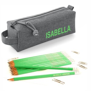 Peut inclure: Une trousse à crayons grise avec une fermeture éclair et une poignée. La trousse porte le nom "ISABELLA" imprimé en vert. La trousse est ouverte et contient 12 crayons verts avec le nom "ISABELLA HOWDEN" imprimé en or. Il y a aussi plusieurs trombones argentés.