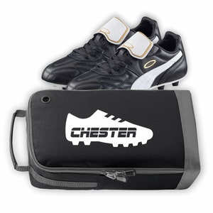 Puede incluir: Botas de fútbol negras y blancas con un acento dorado en la correa. Las botas están en una bolsa de zapatos negra y gris con un contorno blanco de una bota de fútbol y la palabra "CHESTER" impresa en ella.