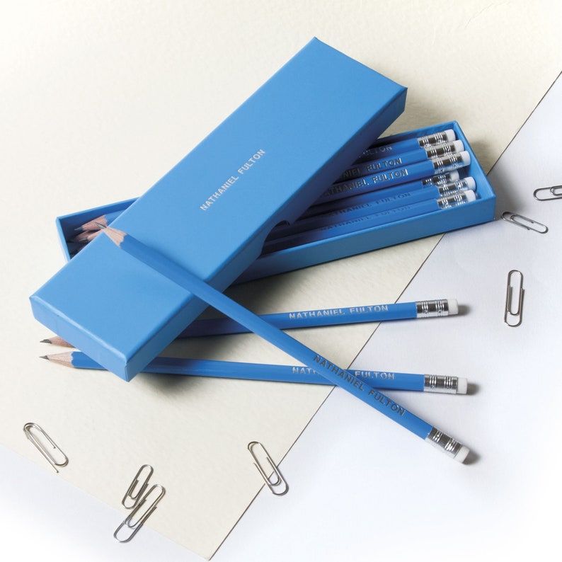 May include: A blue box with the text "Nathaniel Fulton" on the top, containing a set of blue pencils with the same text printed on them. The pencils are arranged in the box and some are sticking out. There are also some paper clips on a white surface.