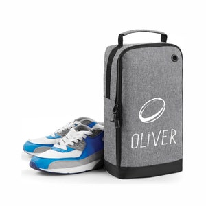 Könnte beinhalten: Graue Schuh Tasche mit einem schwarzen Reißverschluss und einem schwarzen Griff. Die Tasche hat ein weißes Oval mit einer weißen Linie durch und den Namen "OLIVER" darauf gedruckt. Ein Paar blaue und weiße Turnschuhe stehen vor der Tasche.