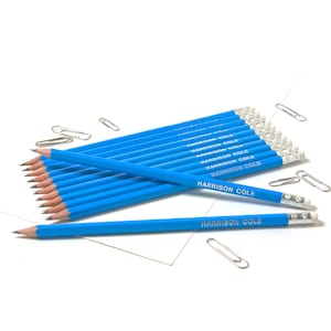 Peut inclure: Un ensemble de 10 crayons bleus avec le nom "HARRISON COLE" imprimé en argent sur chaque crayon. Les crayons sont disposés en éventail sur une surface blanche.
