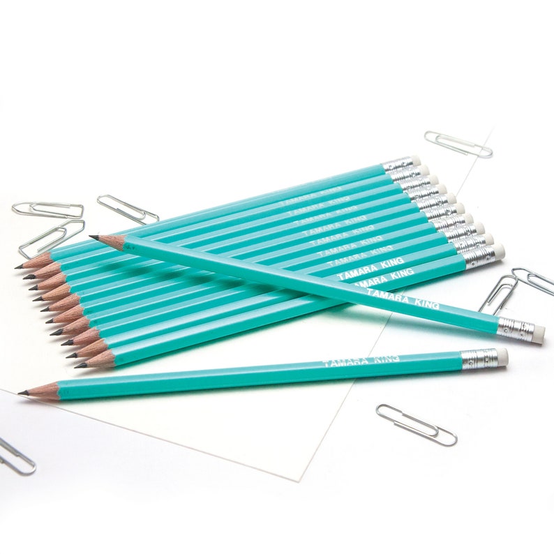 May include: A stack of turquoise blue pencils with white lettering that reads "TAMARA KING" on each pencil. The pencils are arranged on a white surface with silver paper clips scattered around.