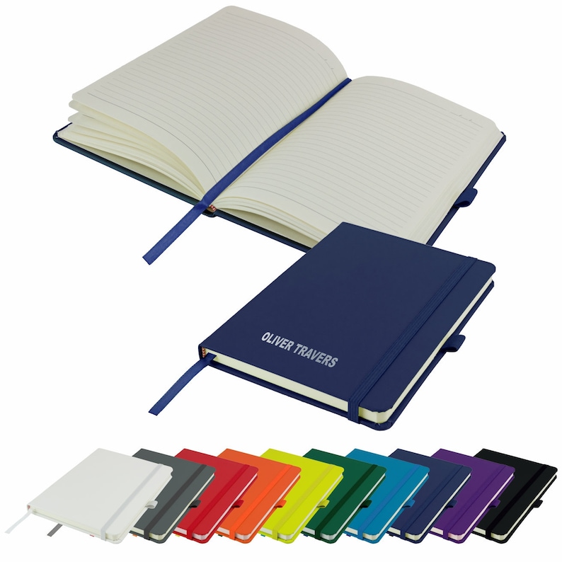 May include: A blue leather-bound notebook with a ribbon bookmark and a personalized inscription that reads "Oliver Travers". The notebook is closed and lying on a white surface. A row of 12 notebooks in various colors is shown below the blue notebook.