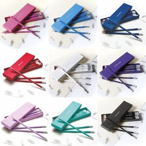 May include: Assortment of ten pencil boxes in various colors, including pink, blue, white, red, purple, teal, and black. Each box contains a set of pencils with the words "Write & Shine" printed on them.