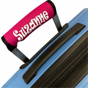 Puede incluir: Una funda de mango de equipaje rosa con el nombre "Suzanne" impreso en letras blancas.