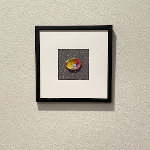 May include: A framed artwork featuring a small, colorful, painted seashell with a gold rim on a gray background with gold polka dots.