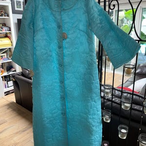 May include: A bright turquoise quilted robe featuring a round neckline, three-quarter length sleeves, and a full-length front button closure. The fabric displays an intricate, tonal embossed pattern of fish and floral motifs throughout the garment.