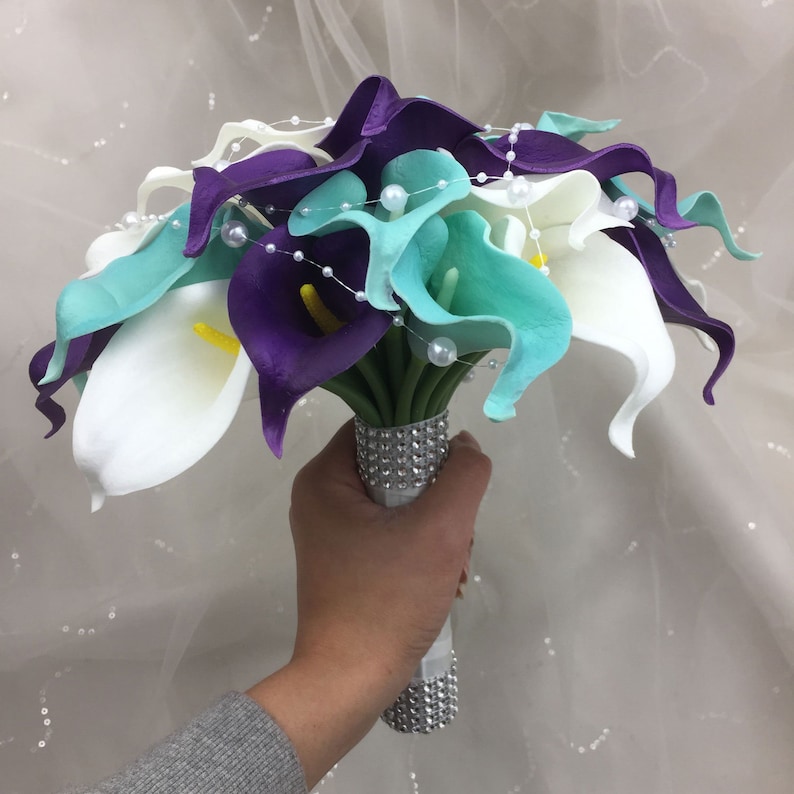 May include: A bridal bouquet of white, teal, and purple calla lilies with pearl accents. The bouquet is wrapped in a silver sparkly ribbon.