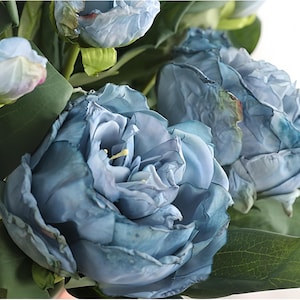 May include: Close-up of a bouquet of blue artificial flowers. The flowers have multiple layers of petals and are surrounded by green leaves. The color of the flowers is a muted blue with darker shades in the center.