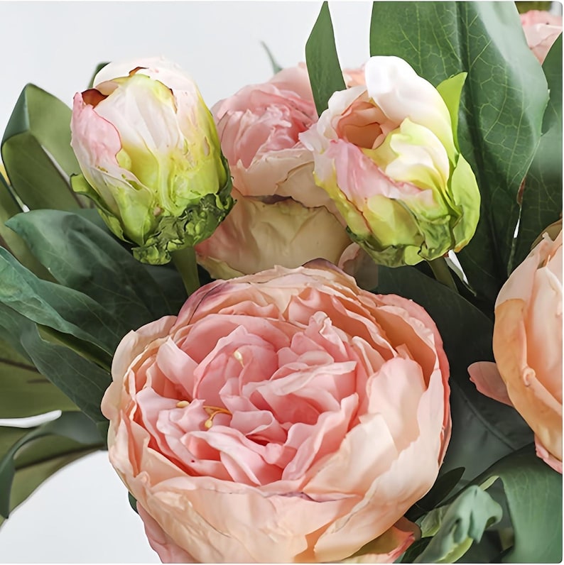 May include: Close-up of a bouquet of artificial peonies. The flowers are in various stages of bloom, with colors ranging from pale pink to peach. The buds have green and yellow accents, and the leaves are a deep green.
