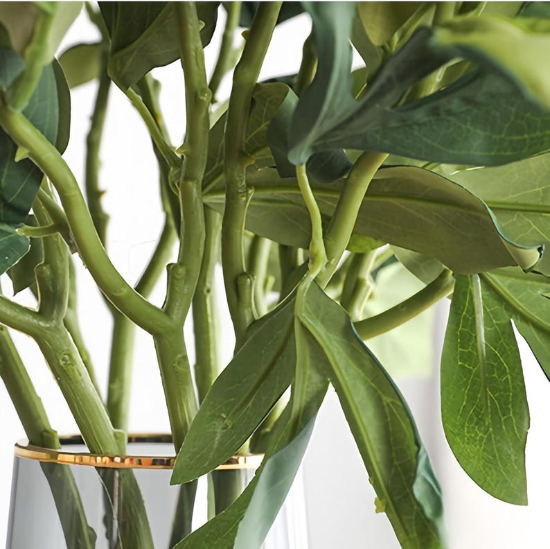 May include: Close-up of a vase filled with artificial green leaves and stems. The leaves have a natural appearance with detailed veining. The vase is clear with a gold rim, adding a touch of elegance to the floral arrangement.