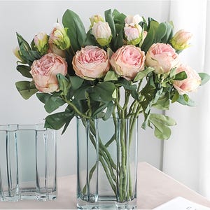 May include: A bouquet of pink peonies and green leaves in a tall, rectangular, light blue glass vase. The flowers are in full bloom, with some buds still closed. A smaller, matching vase sits to the left. The arrangement is on a light pink surface.