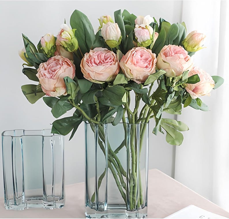 May include: A bouquet of pink peonies and green leaves in a tall, rectangular, light blue glass vase. The flowers are in full bloom, with some buds still closed. A smaller, matching vase sits to the left. The arrangement is on a light pink surface.