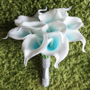 May include: A bouquet of white calla lilies with blue centers. The lilies are arranged in a cluster and tied together with a silver ribbon and rhinestones.