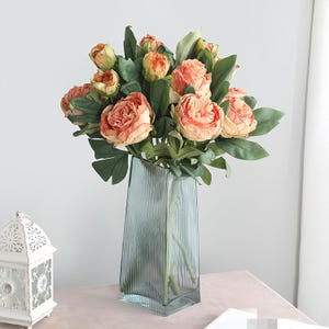 May include: A bouquet of peach-colored roses and green leaves in a tall, blue-green, ribbed glass vase. The flowers are in full bloom, with multiple layers of petals. The vase is rectangular with a slightly angled top. The arrangement sits on a pink surface.