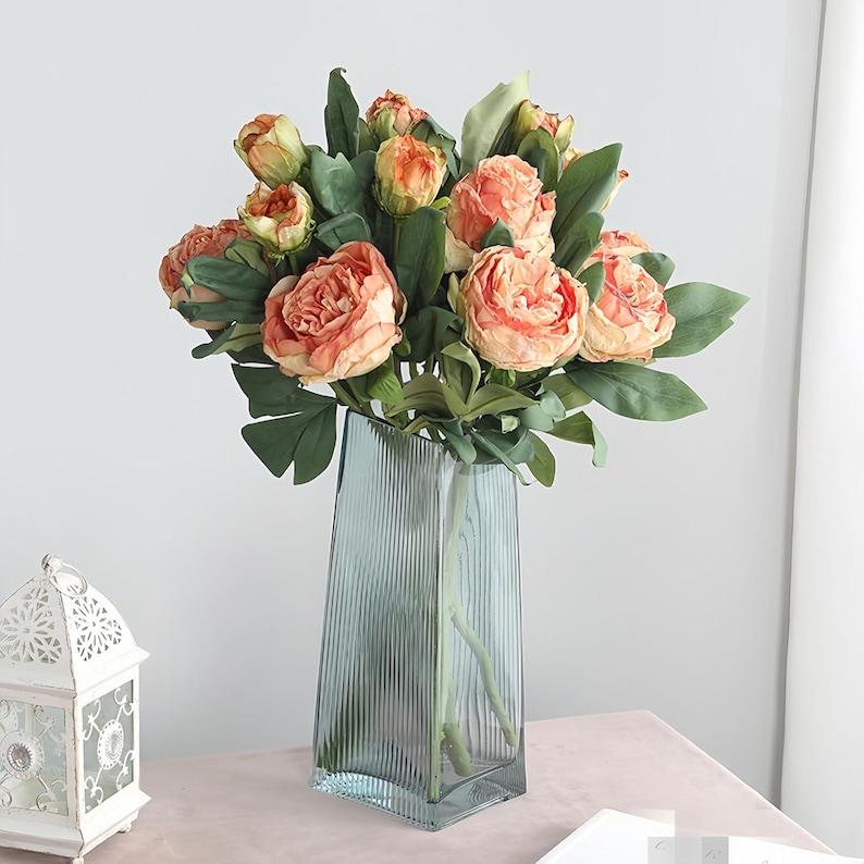 May include: A bouquet of peach-colored roses and green leaves in a tall, blue-green, ribbed glass vase. The flowers are in full bloom, with multiple layers of petals. The vase is rectangular with a slightly angled top. The arrangement sits on a pink surface.