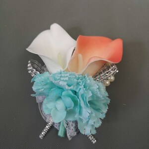 May include: A wedding boutonniere with a white and peach calla lily, a light blue carnation, and silver and pearl embellishments.