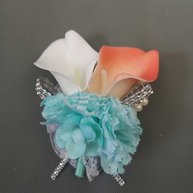 May include: A wedding boutonniere with a white and peach calla lily, a light blue carnation, and silver and pearl embellishments.