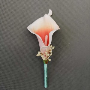 May include: A single peach and white artificial calla lily flower boutonniere with a teal stem and small white flower accents.