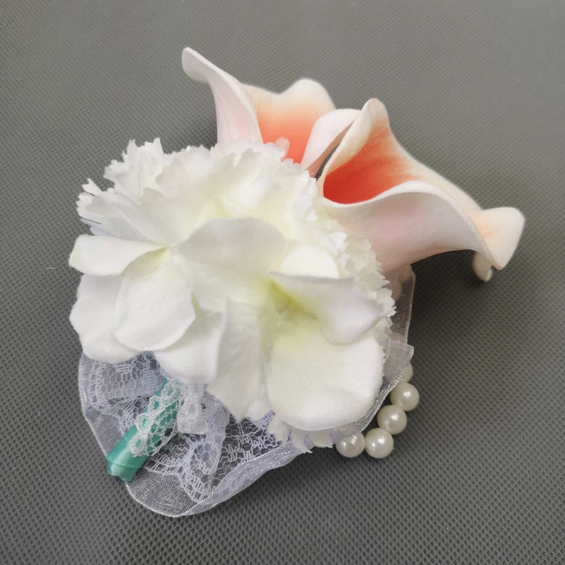 May include: A white and peach artificial flower boutonniere with a lace and pearl trim. The boutonniere has a teal ribbon.