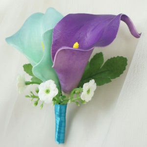 May include: A wedding boutonniere with two calla lilies, one light blue and one purple, with white baby's breath and a teal ribbon.