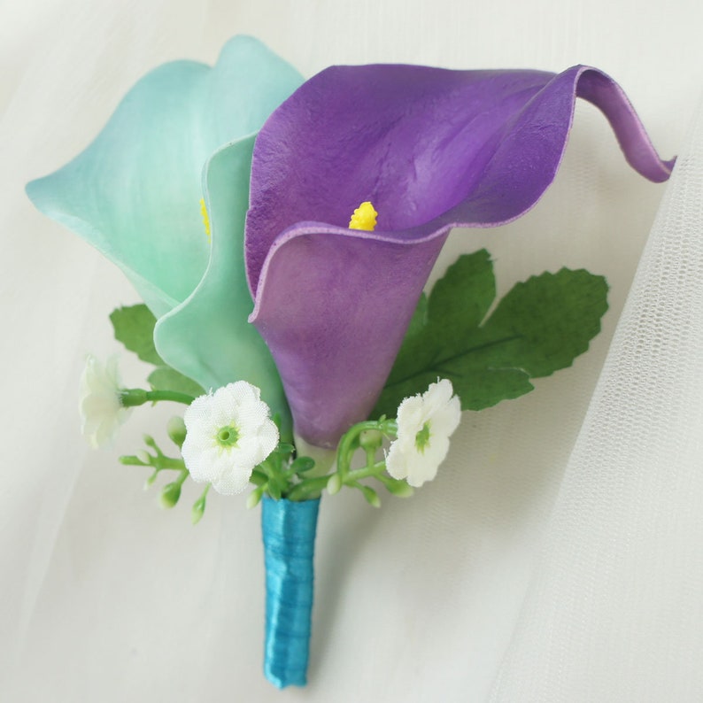 May include: A wedding boutonniere with two calla lilies, one light blue and one purple, with white baby's breath and a teal ribbon.