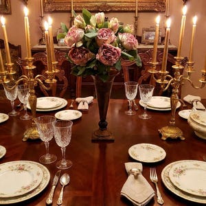May include: A formal dining table set for a meal. The table is made of dark wood and is set with floral patterned plates, crystal glasses, and gold candle holders with lit candles. A vase of pink flowers sits in the center.