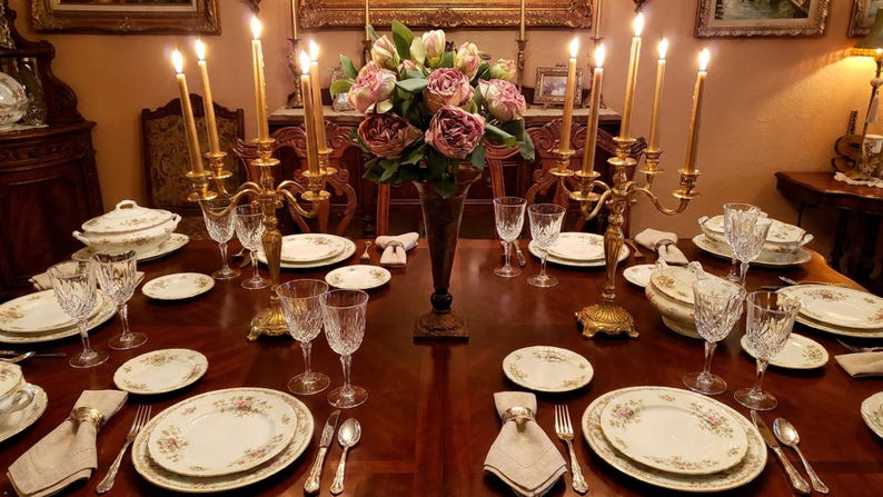 May include: A formal dining table set for a meal. The table is made of dark wood and is set with floral patterned plates, crystal glasses, and gold candle holders with lit candles. A vase of pink flowers sits in the center.