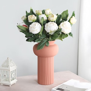 May include: A peach-colored vase with a round top and a ribbed base holds a bouquet of white peonies and green leaves. A white decorative lantern is on the left. A magazine is on the right.