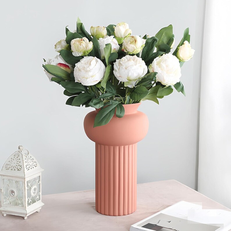May include: A peach-colored vase with a round top and a ribbed base holds a bouquet of white peonies and green leaves. A white decorative lantern is on the left. A magazine is on the right.