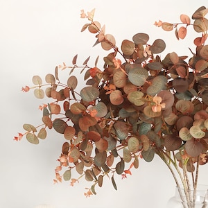 May include: A bouquet of artificial eucalyptus branches with round, muted green and brown leaves. The stems are a light brown color, and small, peach-colored buds are scattered throughout the arrangement. The leaves have a slight sheen.
