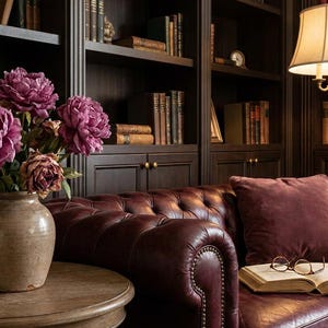 May include: A cozy library scene featuring a burgundy leather sofa with a matching pillow and a round wooden side table. A vase of purple peonies sits on the table. Bookshelves and a floor lamp complete the room.