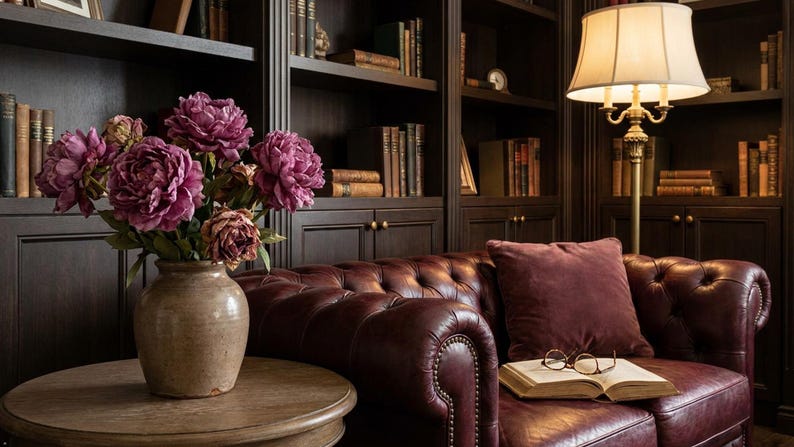 May include: A cozy library scene featuring a burgundy leather sofa with a matching pillow and a round wooden side table. A vase of purple peonies sits on the table. Bookshelves and a floor lamp complete the room.