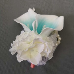 May include: A white and teal artificial flower arrangement with a white lace and pearl embellishment. The arrangement features a calla lily and a carnation.