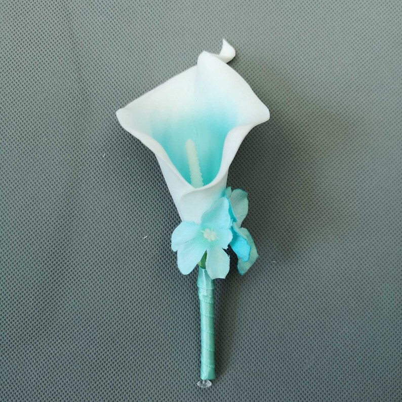 May include: A white and teal artificial calla lily boutonniere with teal accent flowers. The boutonniere is attached to a teal ribbon.