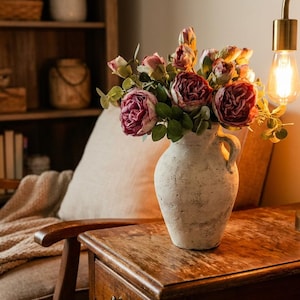 May include: A rustic, off-white vase filled with burgundy and pink roses sits on a wooden side table. A gold-toned lamp with a vintage bulb illuminates the scene. A cozy armchair with a beige throw and a wooden bookshelf complete the warm, inviting interior.